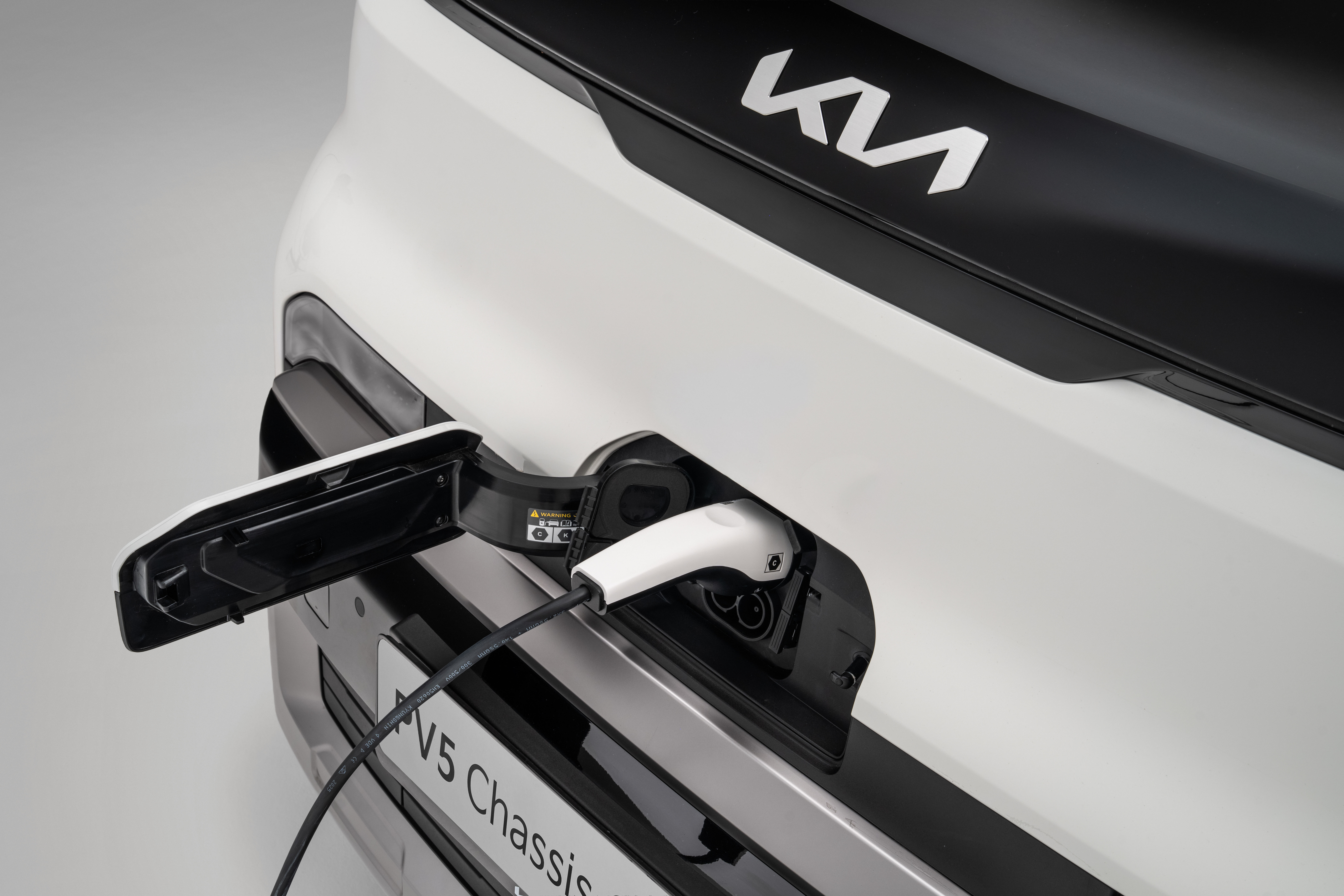 Detalle del puerto de carga trasero del Kia PV5 Chasis Cabina blanco con el cable de carga enchufado y el logo de Kia visible.