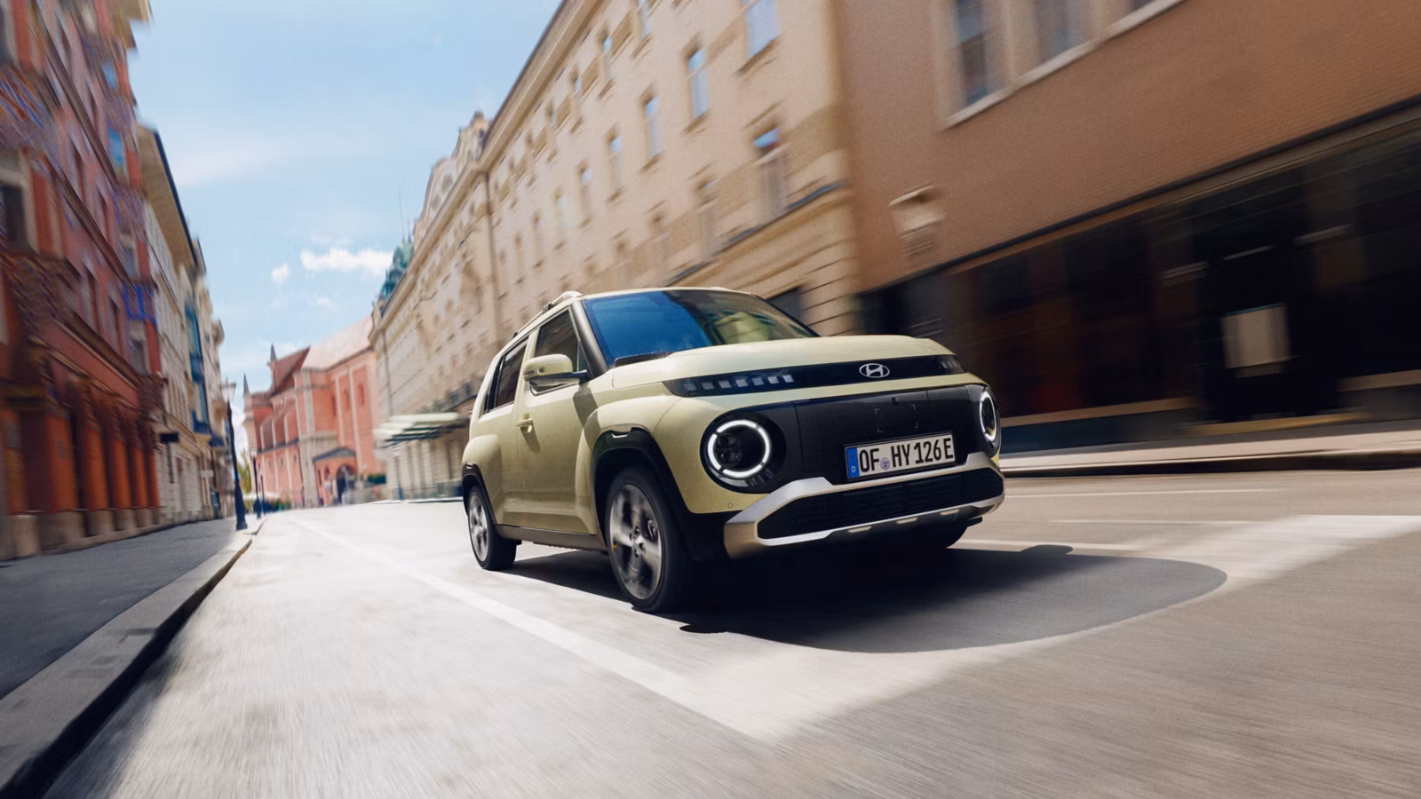 Hyundai Inster beige claro conduciendo por una calle urbana empedrada con edificios históricos, con efecto de movimiento