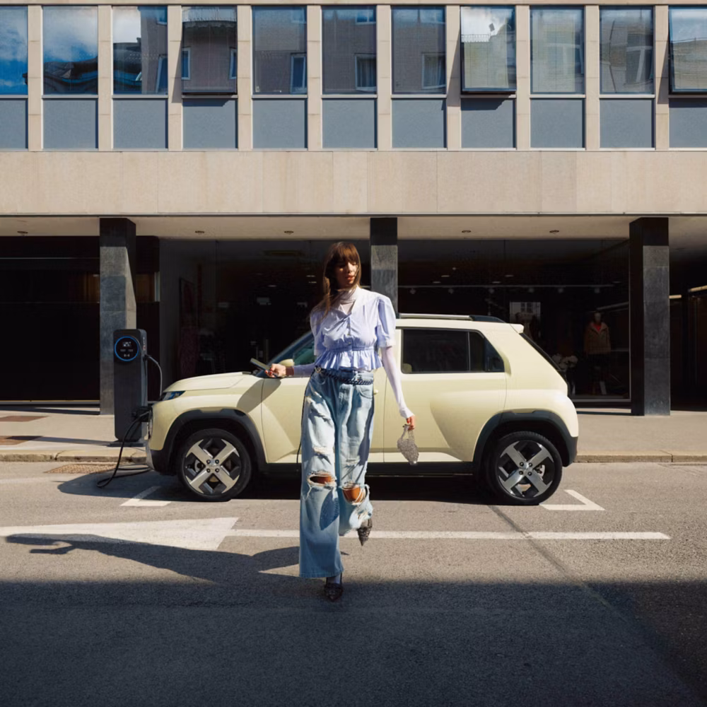 Hyundai Inster beige claro cargando en la calle junto a un edificio moderno, con una persona de pie delante