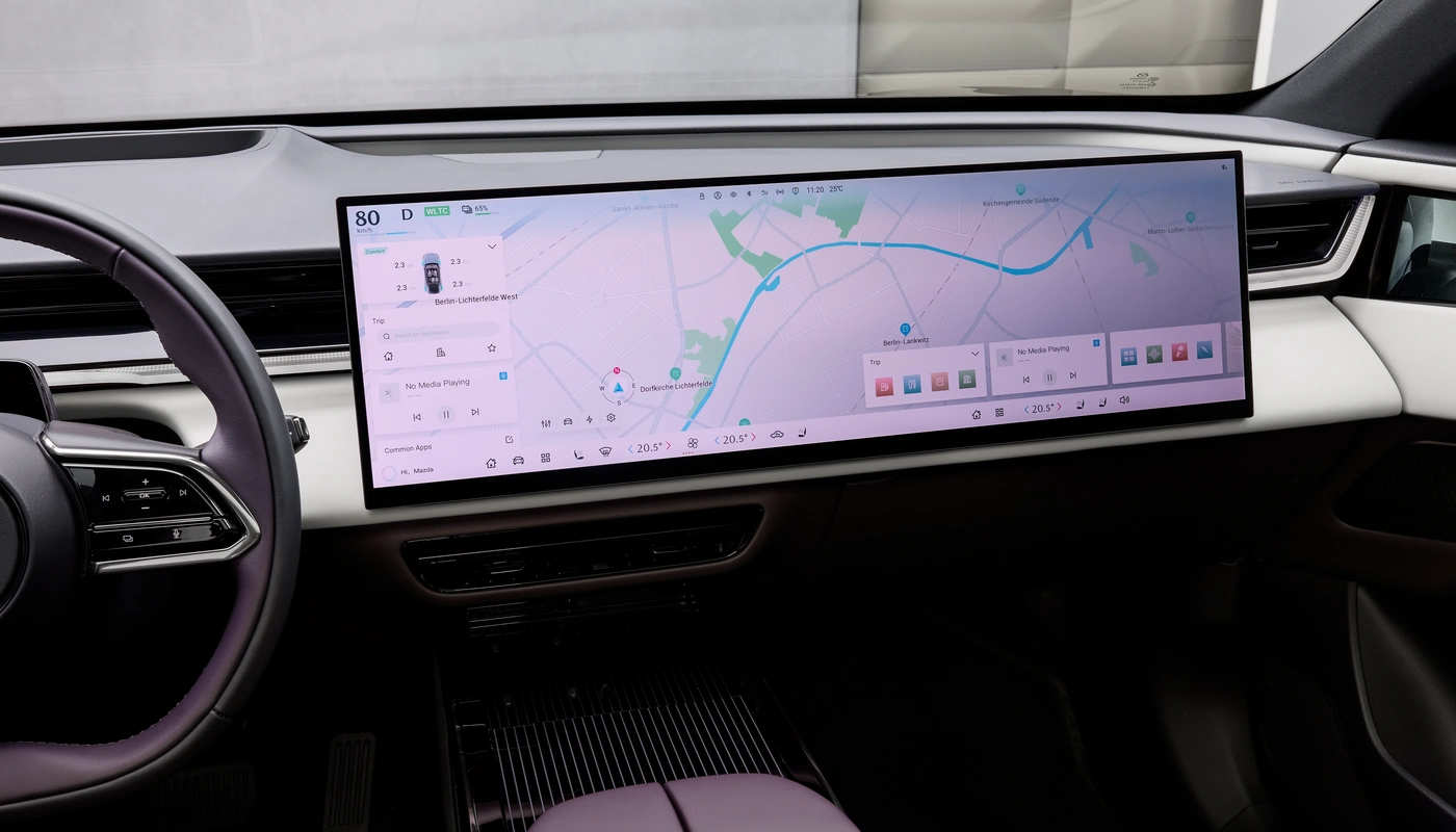 Interior del CX-6e con el volante con logo Mazda y la gran pantalla central mostrando una interfaz con un modelo 3D del coche.