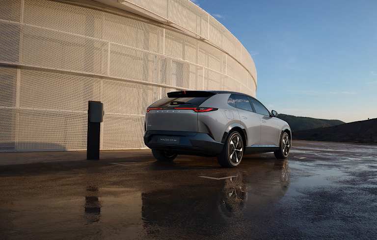 Mazda CX-6e gris claro junto a un cargador eléctrico con un edificio moderno de fondo.