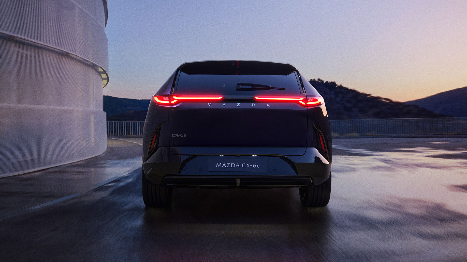 Vista trasera del CX-6e oscuro al atardecer con las luces traseras encendidas, aparcado en una azotea junto a un edificio curvo.