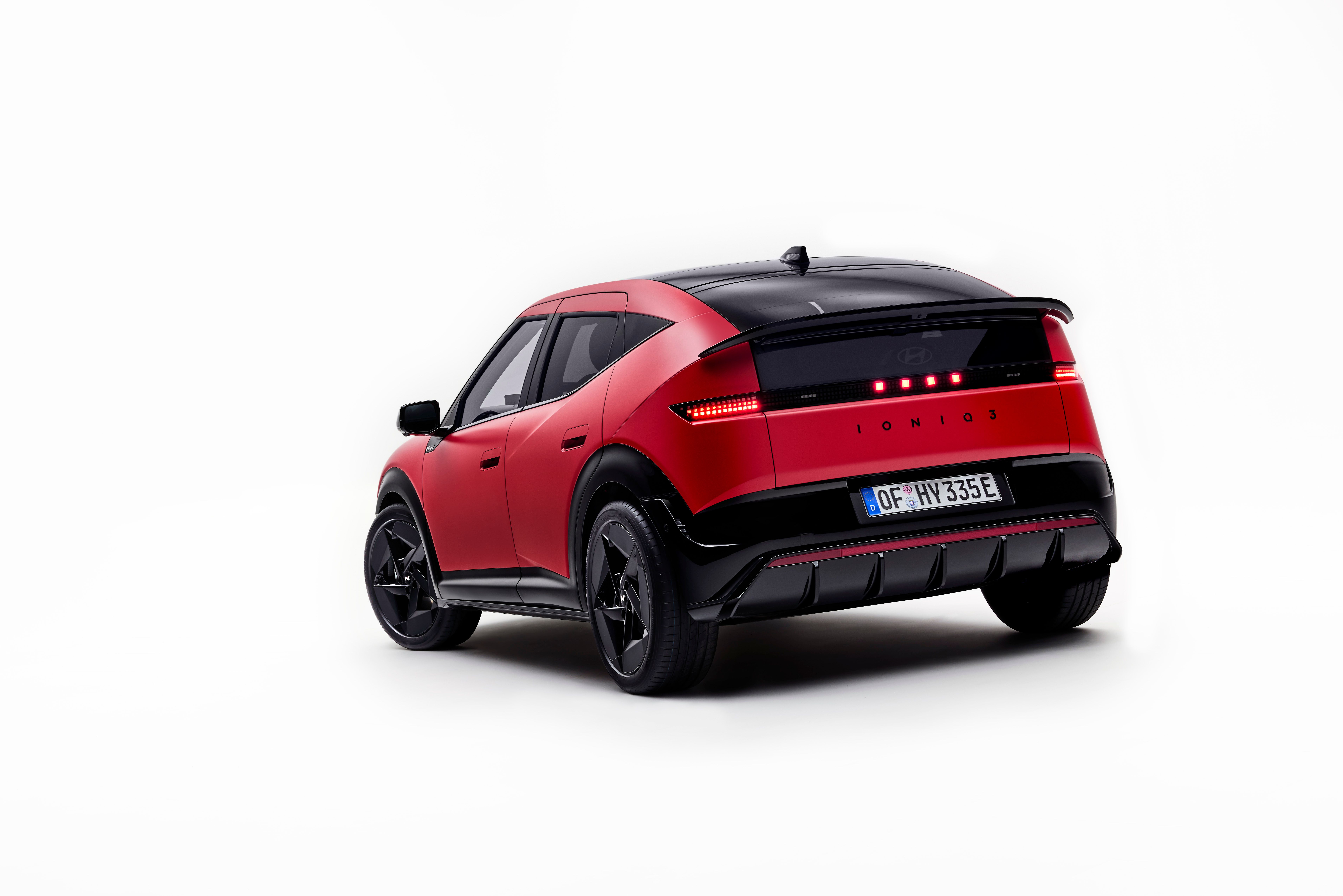 Vista trasera tres cuartos del Hyundai IONIQ 3 rojo con techo negro sobre fondo blanco de estudio, pilotos traseros encendidos y llantas negras de radios múltiples