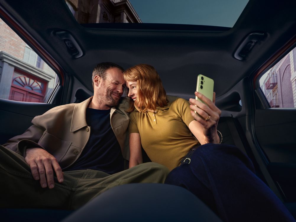Pareja sonriendo y haciéndose un selfie en los asientos traseros bajo el techo panorámico