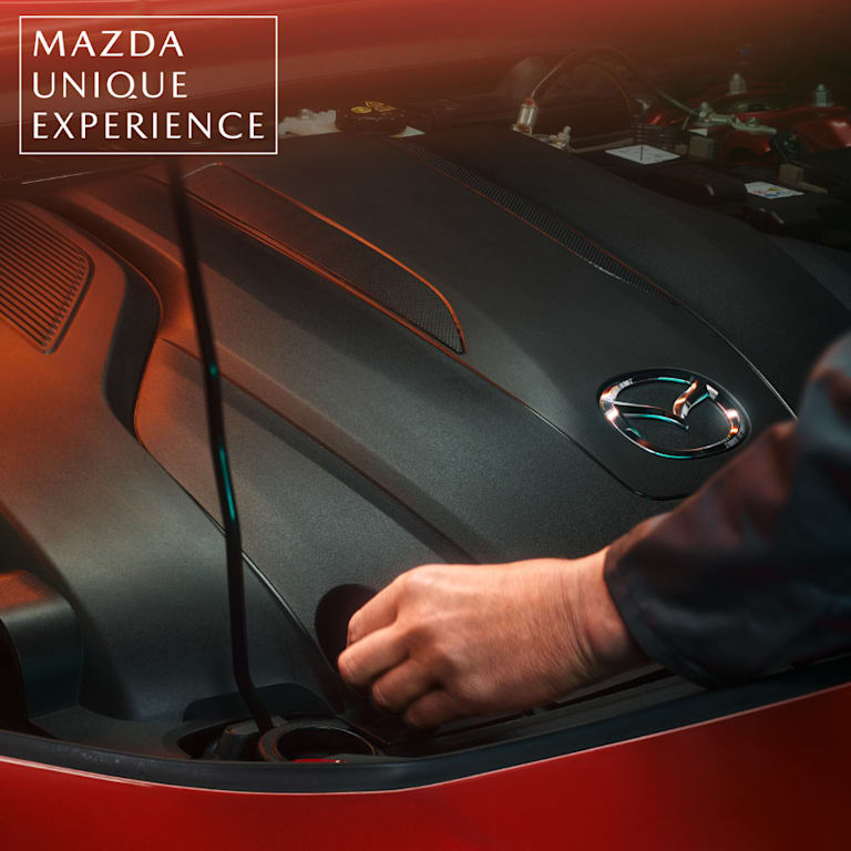 Detalle del motor de un Mazda con el logotipo visible en la cubierta y una mano manipulando un componente