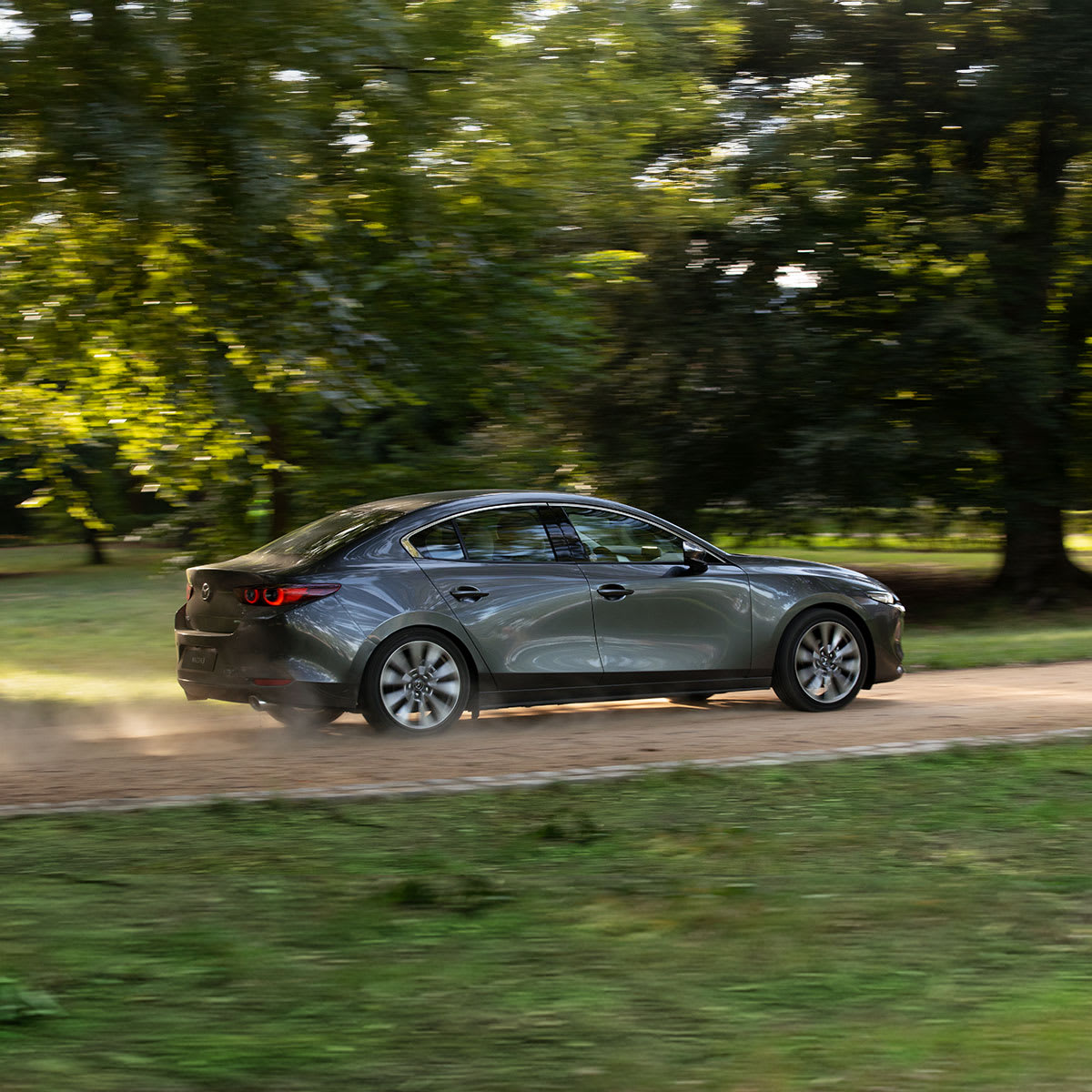 Mazda3 Sedán de color gris oscuro visto de lateral en movimiento sobre un camino, carrocería alternativa de la gama