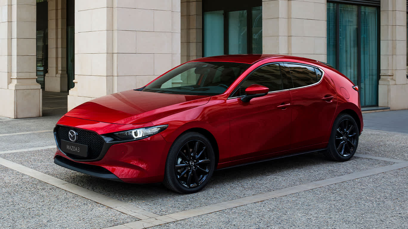Mazda3 rojo aparcado frente a un edificio moderno, visto en tres cuartos frontal