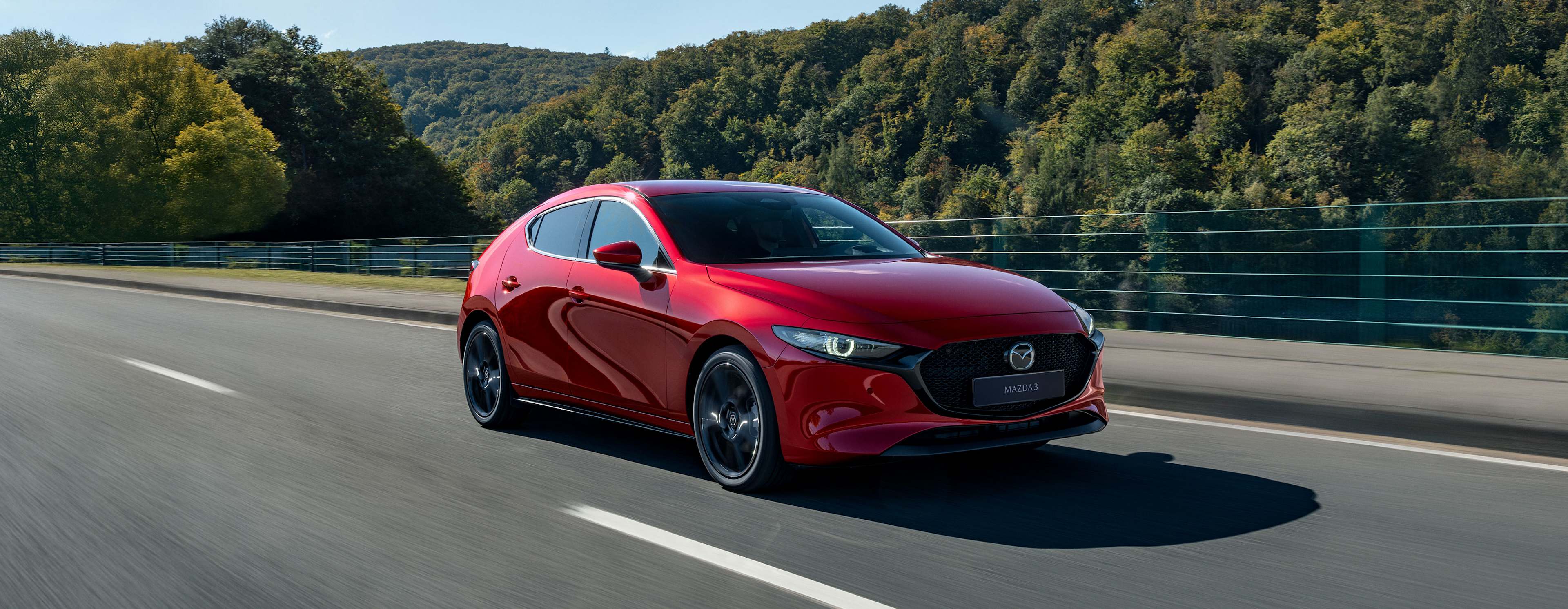Mazda3 rojo hatchback en movimiento sobre una carretera con árboles y cielo azul, en tres cuartos frontal