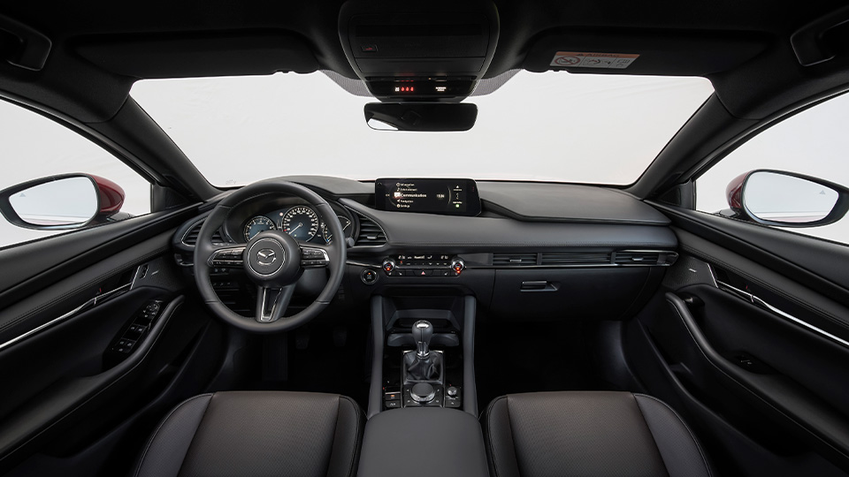 Vista frontal del interior del Mazda3: salpicadero, volante, consola central y asientos delanteros, con los retrovisores reflejando la carrocería roja