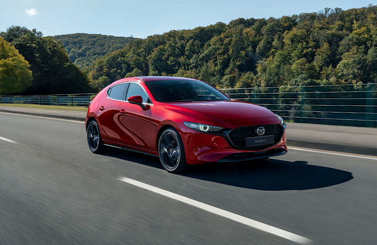 Mazda3 hatchback rojo visto en tres cuartos frontal, en movimiento sobre una carretera con árboles y montañas al fondo