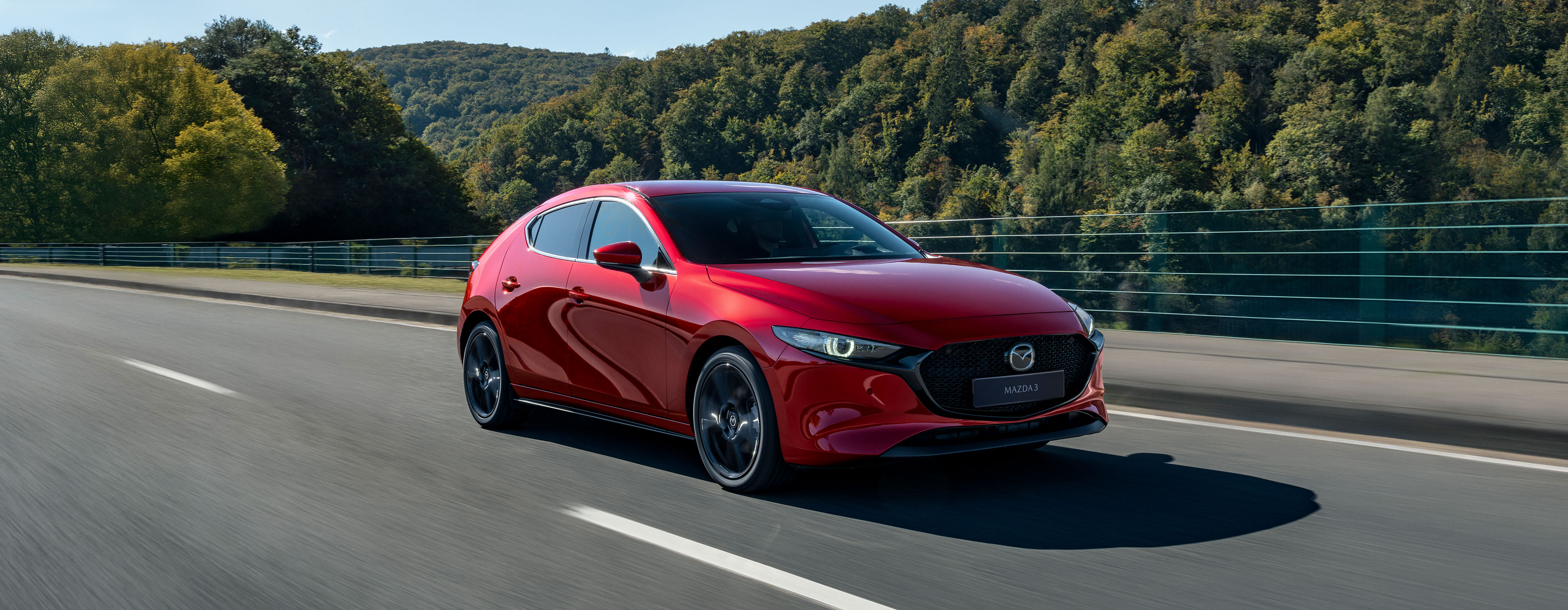 Mazda3 hatchback rojo conduciendo por una carretera entre bosque, en tres cuartos frontal