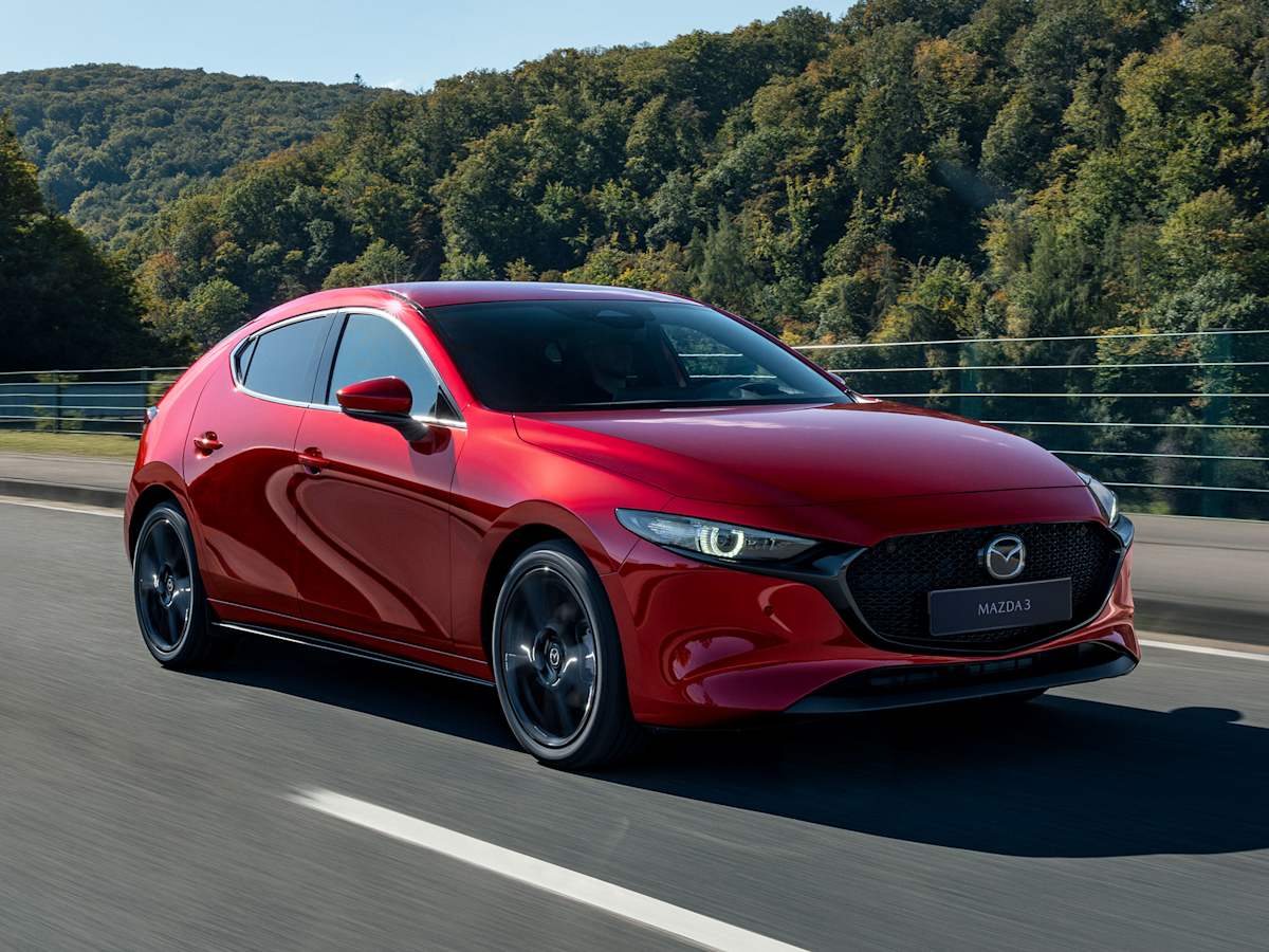 Vista frontal tres cuartos de un Mazda3 rojo circulando por carretera con fondo boscoso, con la luz reflejándose en la carrocería