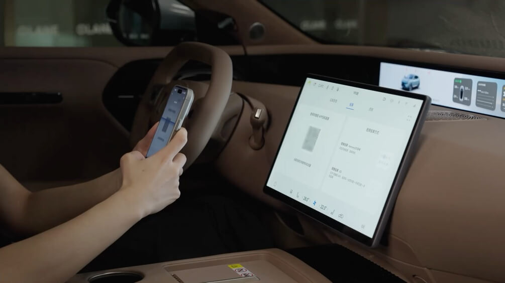 Manos del conductor sosteniendo un teléfono frente a la pantalla central y el cuadro digital del vehículo