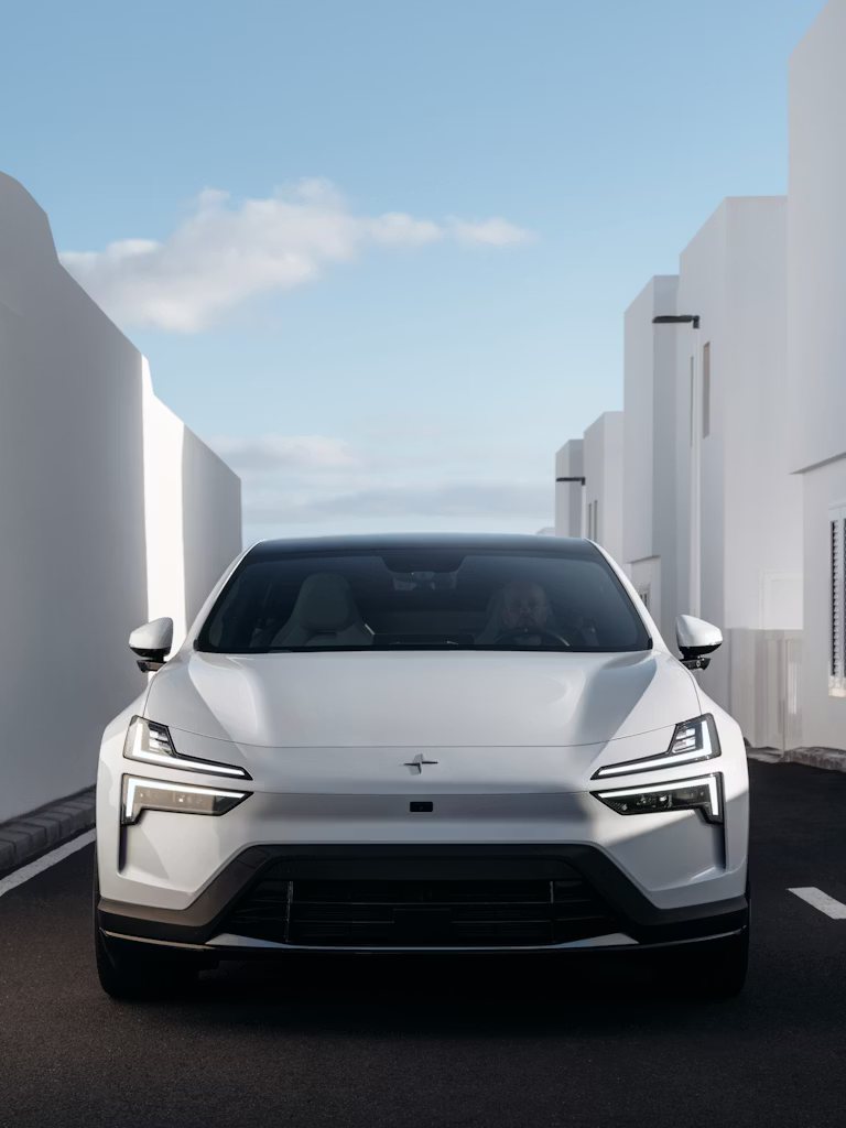 Vista frontal de un Polestar 4 blanco estacionado en una calle de edificios modernos blancos bajo cielo azul.