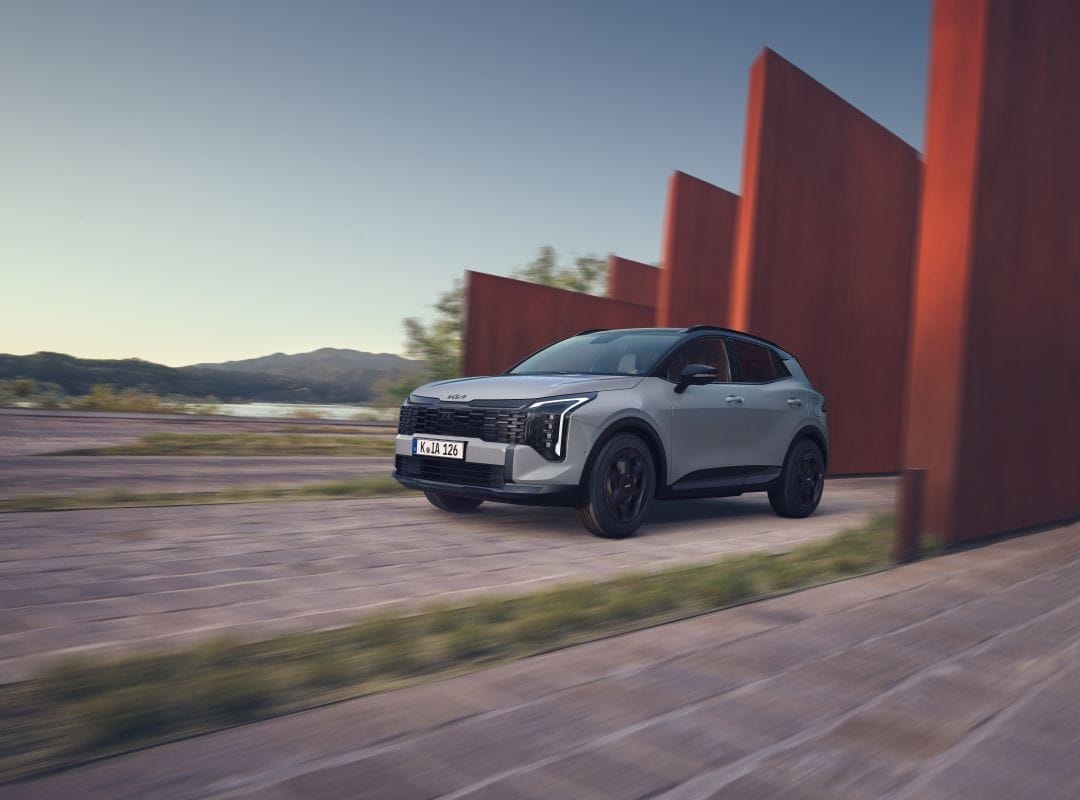 Kia Sportage gris claro circulando por carretera con estructuras rojas y paisaje natural al fondo