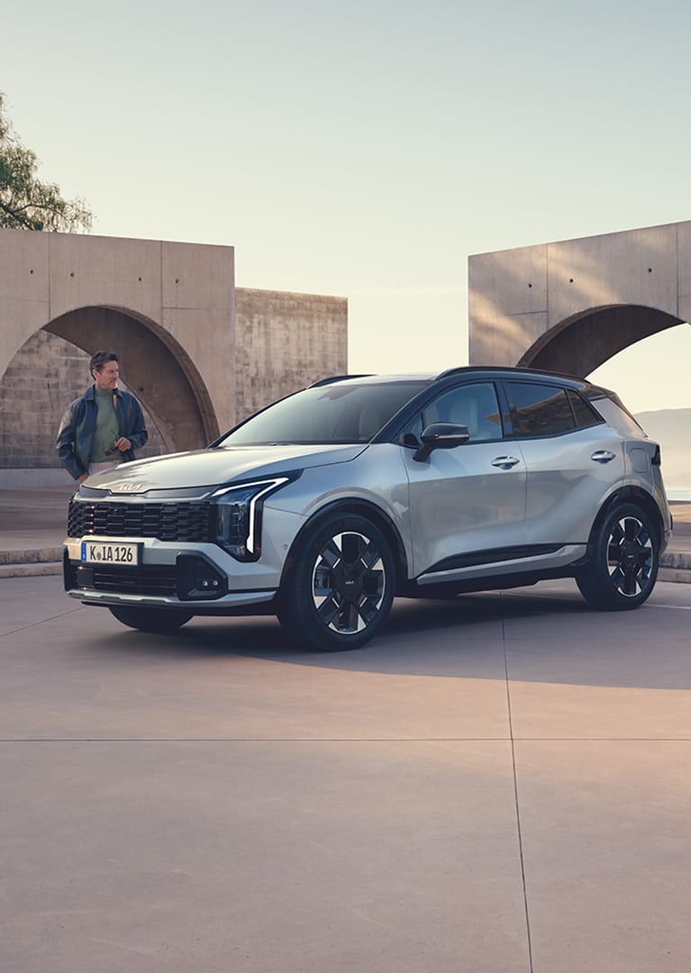 Kia Sportage gris claro con techo negro aparcado frente a una arquitectura moderna con arcos, con un hombre junto al capó