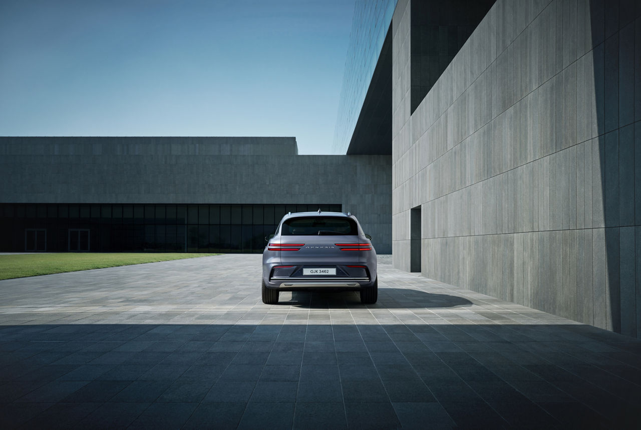 Vista trasera del Electrified GV70 gris oscuro estacionado en una plaza exterior moderna con edificios de hormigón y césped al fondo.