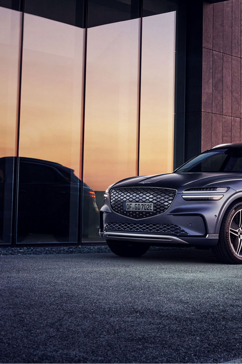 Frontal del Genesis Electrified GV70 gris oscuro con su parrilla cerrada, faros divididos y llantas multirradio bitono, ante un edificio moderno al atardecer.
