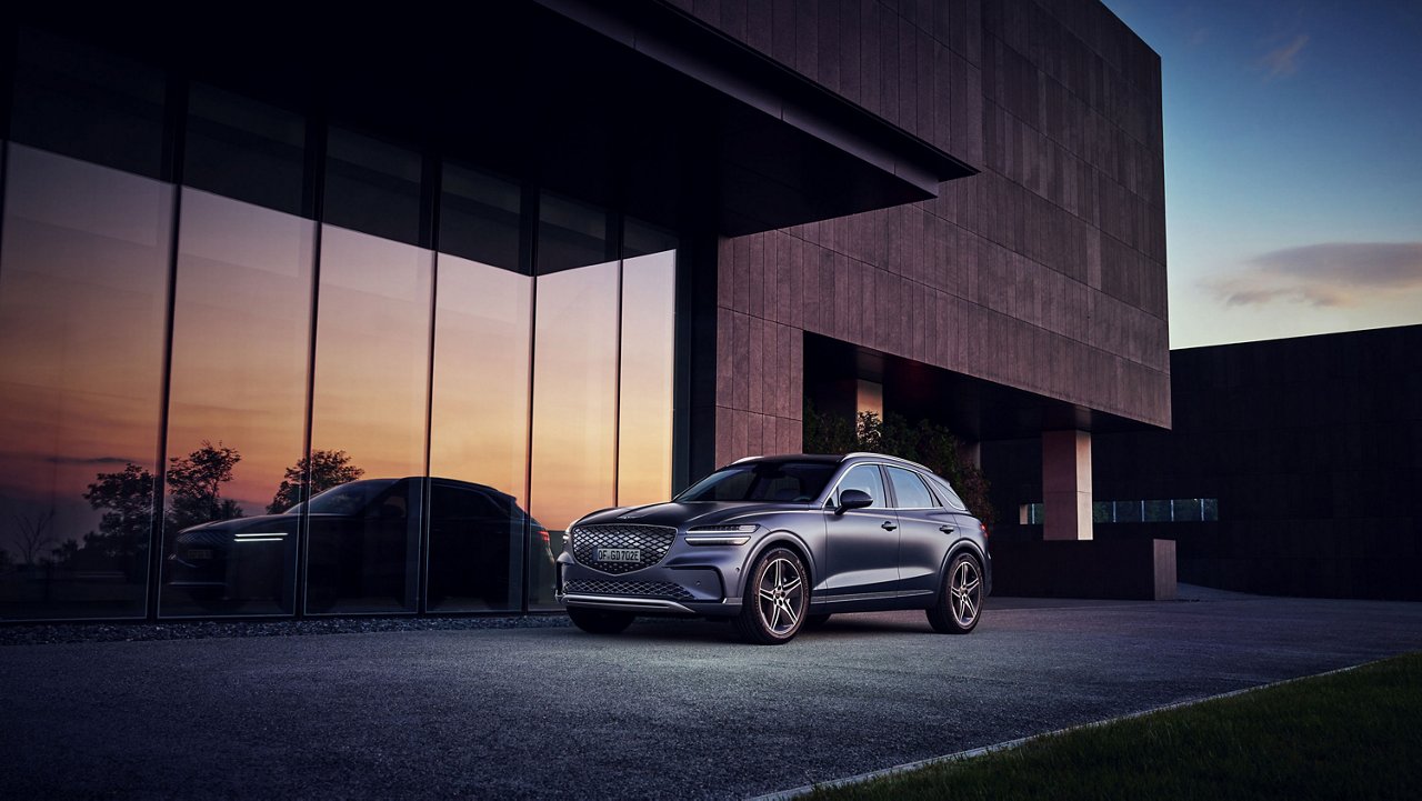 Electrified GV70 gris oscuro aparcado frente a un edificio moderno cuyas ventanas reflejan un atardecer, con iluminación tenue.