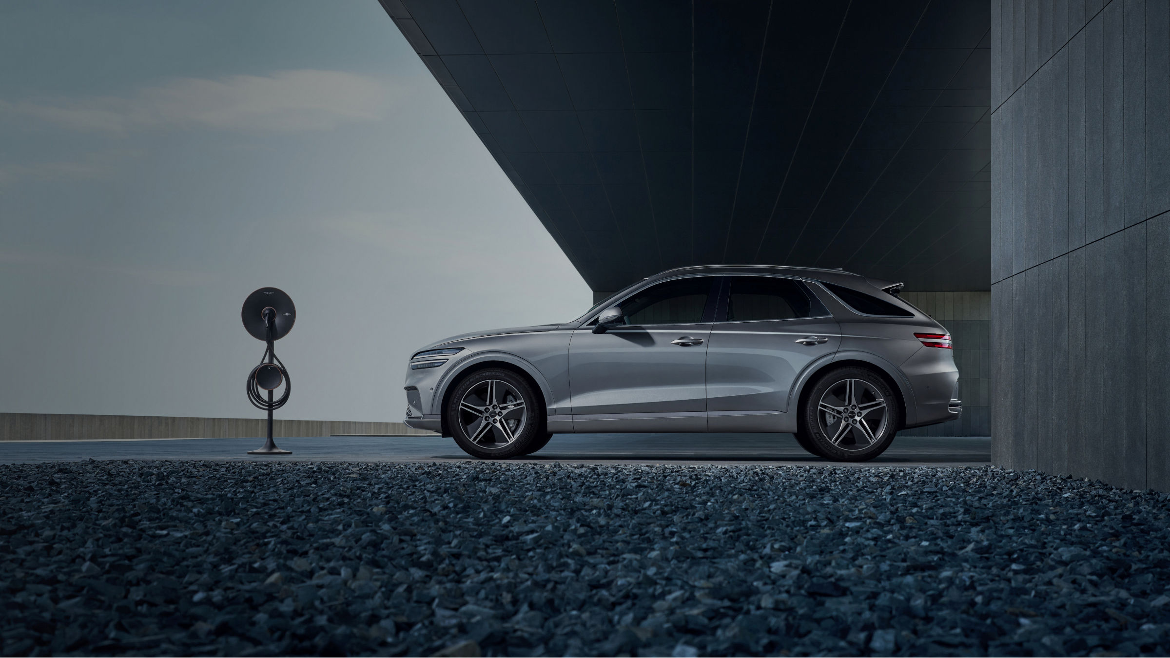 Electrified GV70 gris oscuro estacionado junto a un cargador de vehículo eléctrico, ante una arquitectura moderna de tonos oscuros.