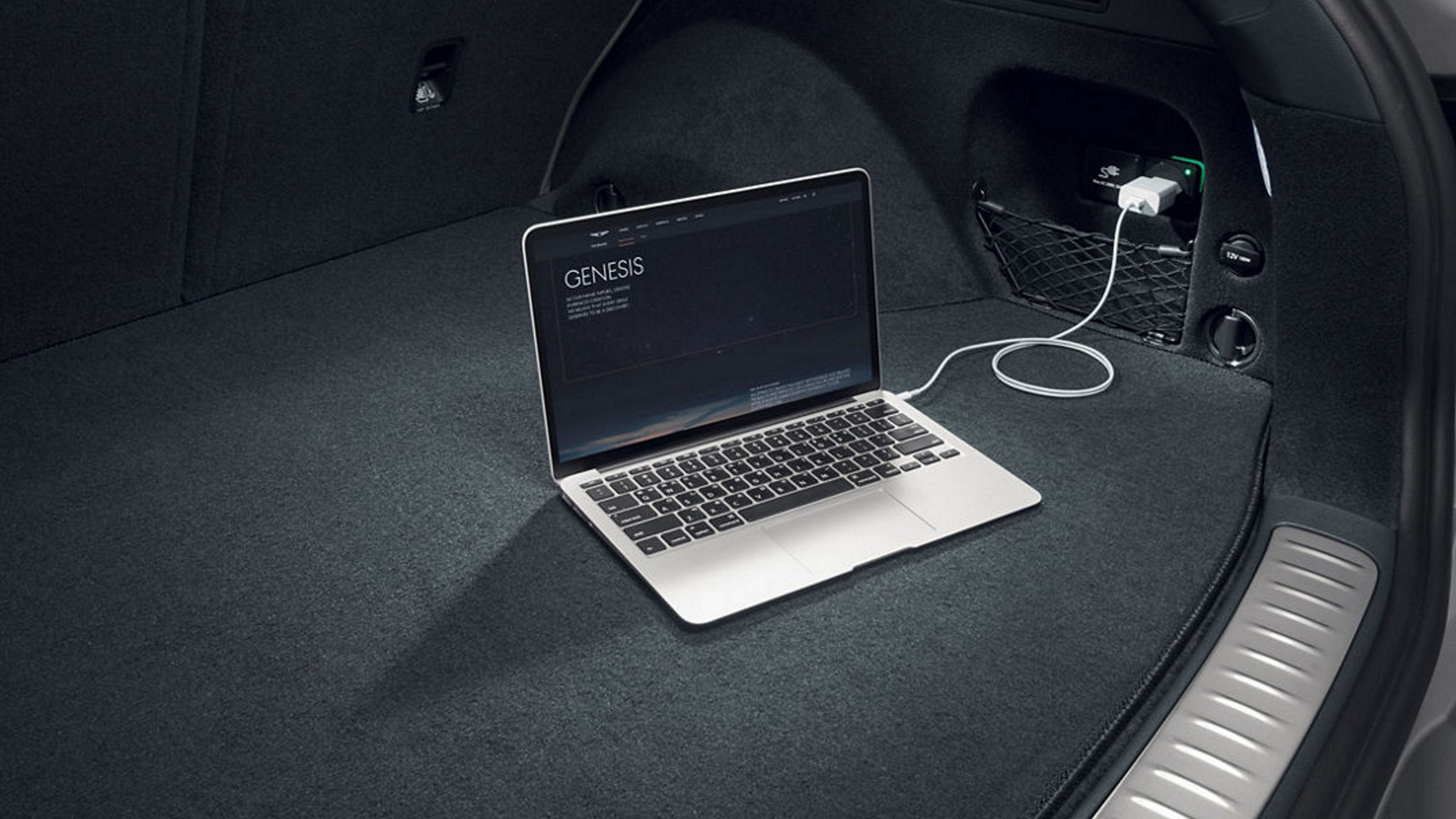 Maletero del vehículo con un portátil conectado a la toma de corriente interior; la pantalla muestra el logotipo Genesis.