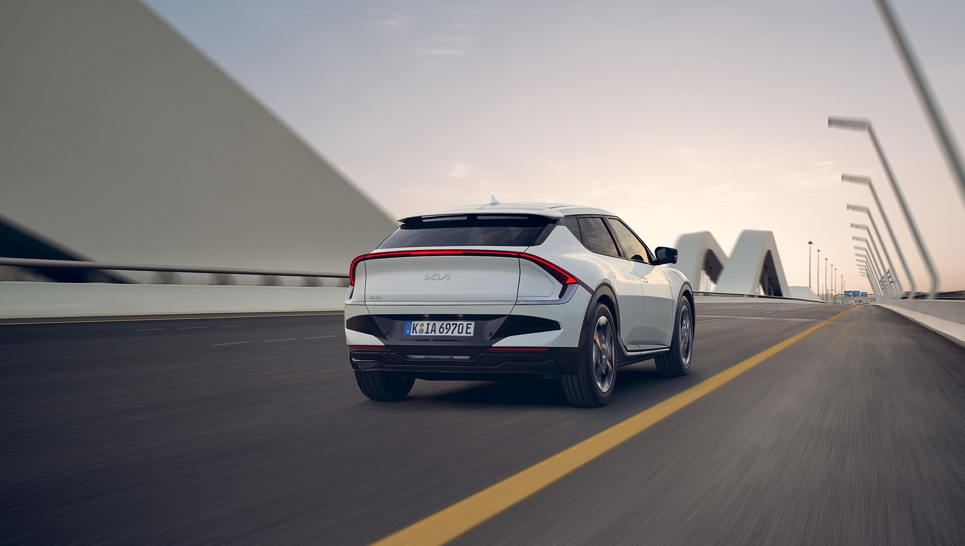 Kia EV6 blanco circulando por un puente al atardecer, en vista trasera tres cuartos