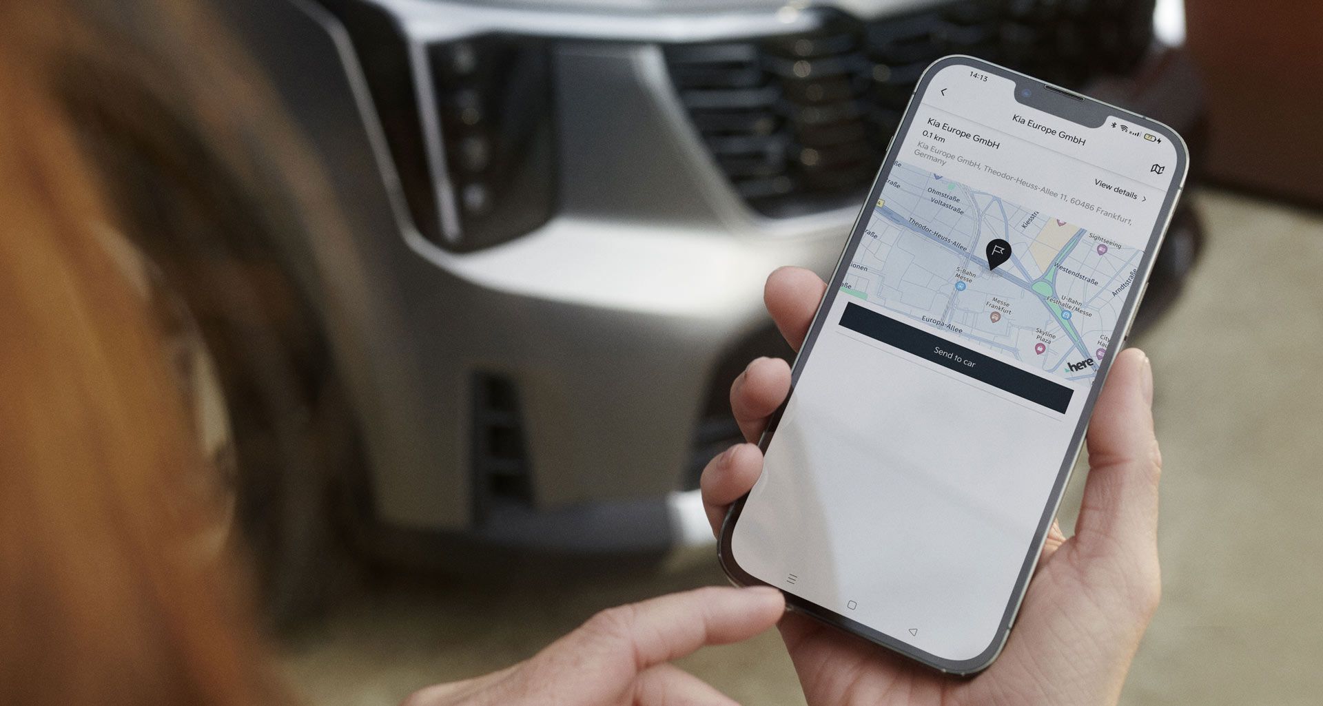 Mano sosteniendo un smartphone con una aplicación de mapas de Kia y un coche desenfocado al fondo
